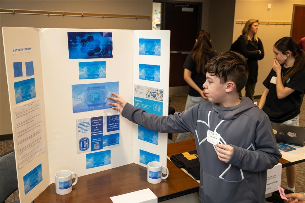  A student is pointing to his tri-fold presentation while explaining his project.